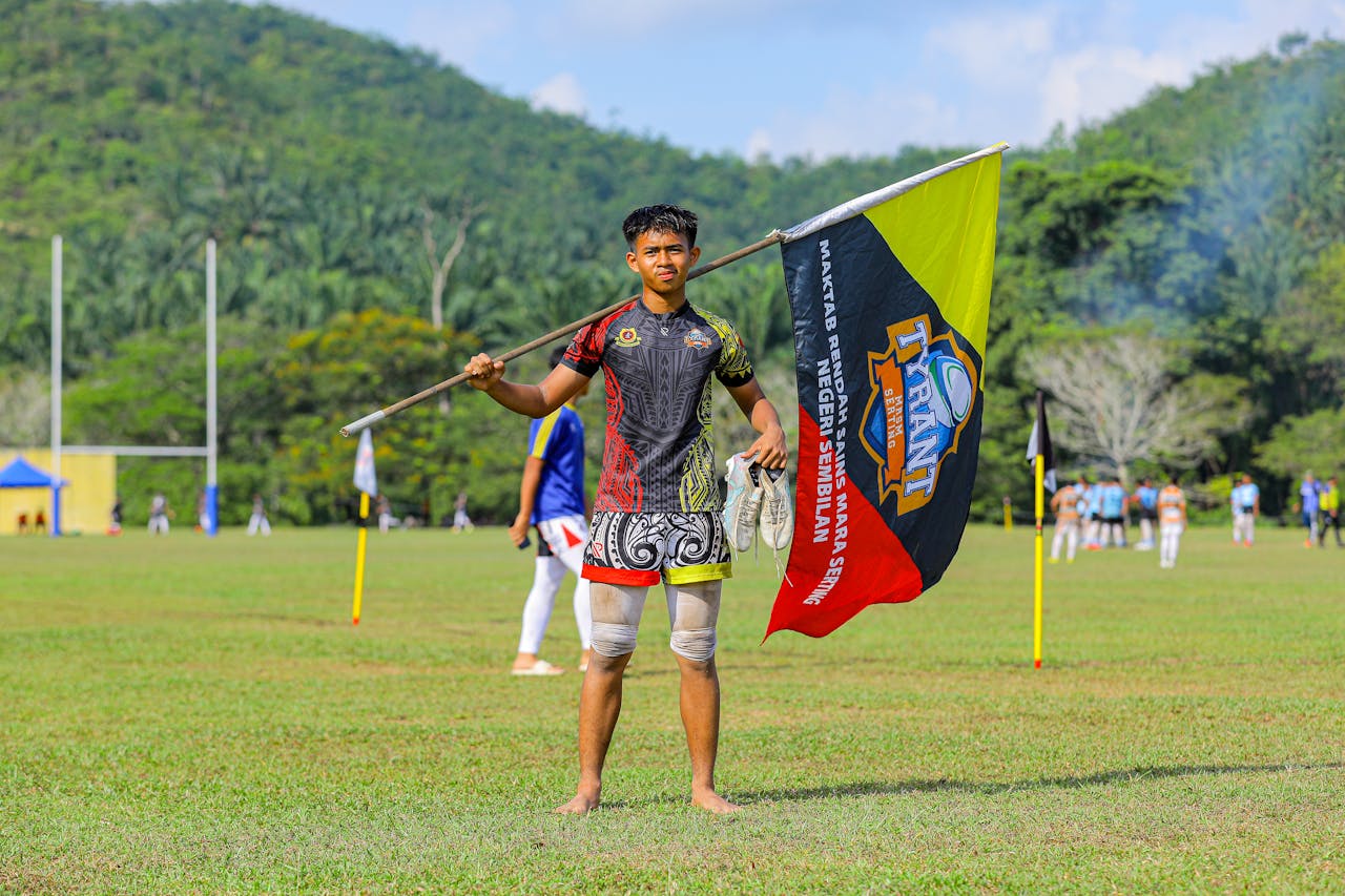 A young athlete holding a flag on an outdoor sports field with hills in the background.